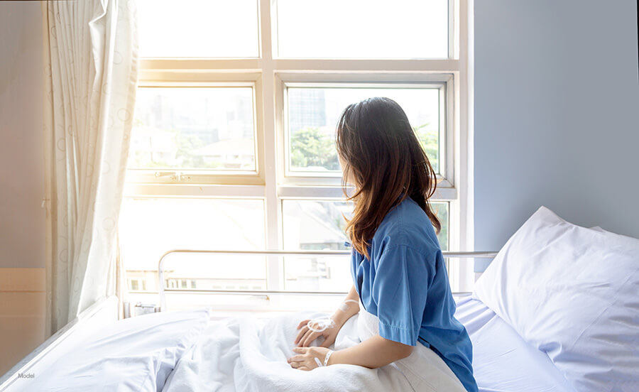 Woman sitting up in hospital bed, looking out a sunny window.